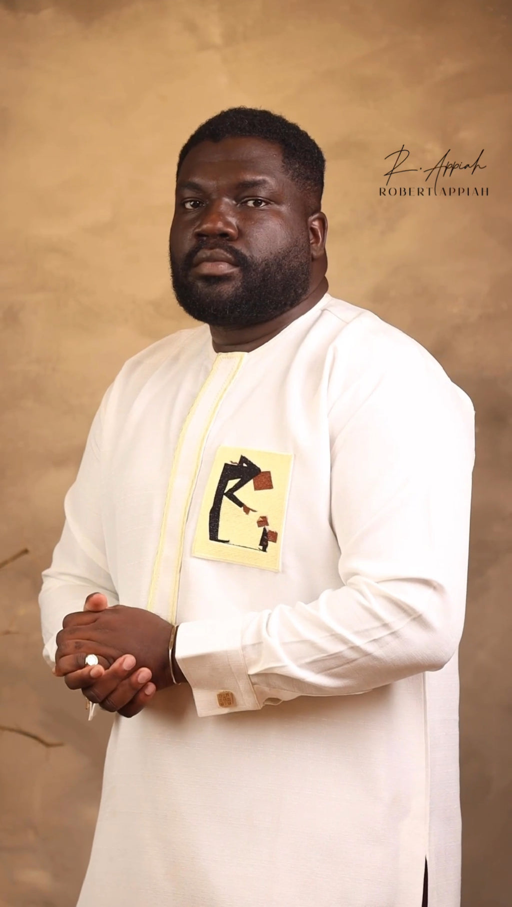 Man in a white traditional outfit with a yellow embroidered design against a beige background